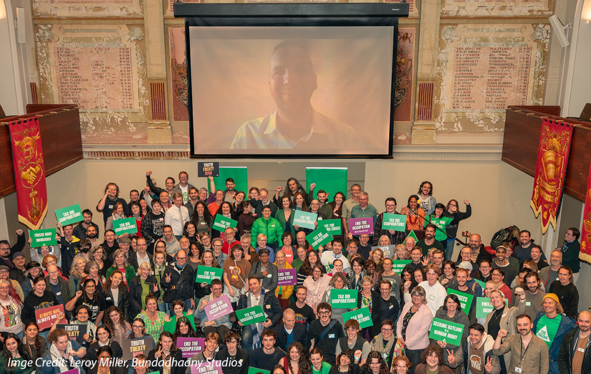 Victorian Greens supporters, members and represenatives at Campaign Con smiling under big screen with Zach Polanski