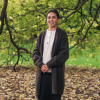 This is an image of Talia, a smiling individual standing outdoors under a big tree. She is wearing a white shirt with a long, dark cardigan on top. The background features a leafy brown ground, branches of a tree curling downwards and lush green grass in the background.