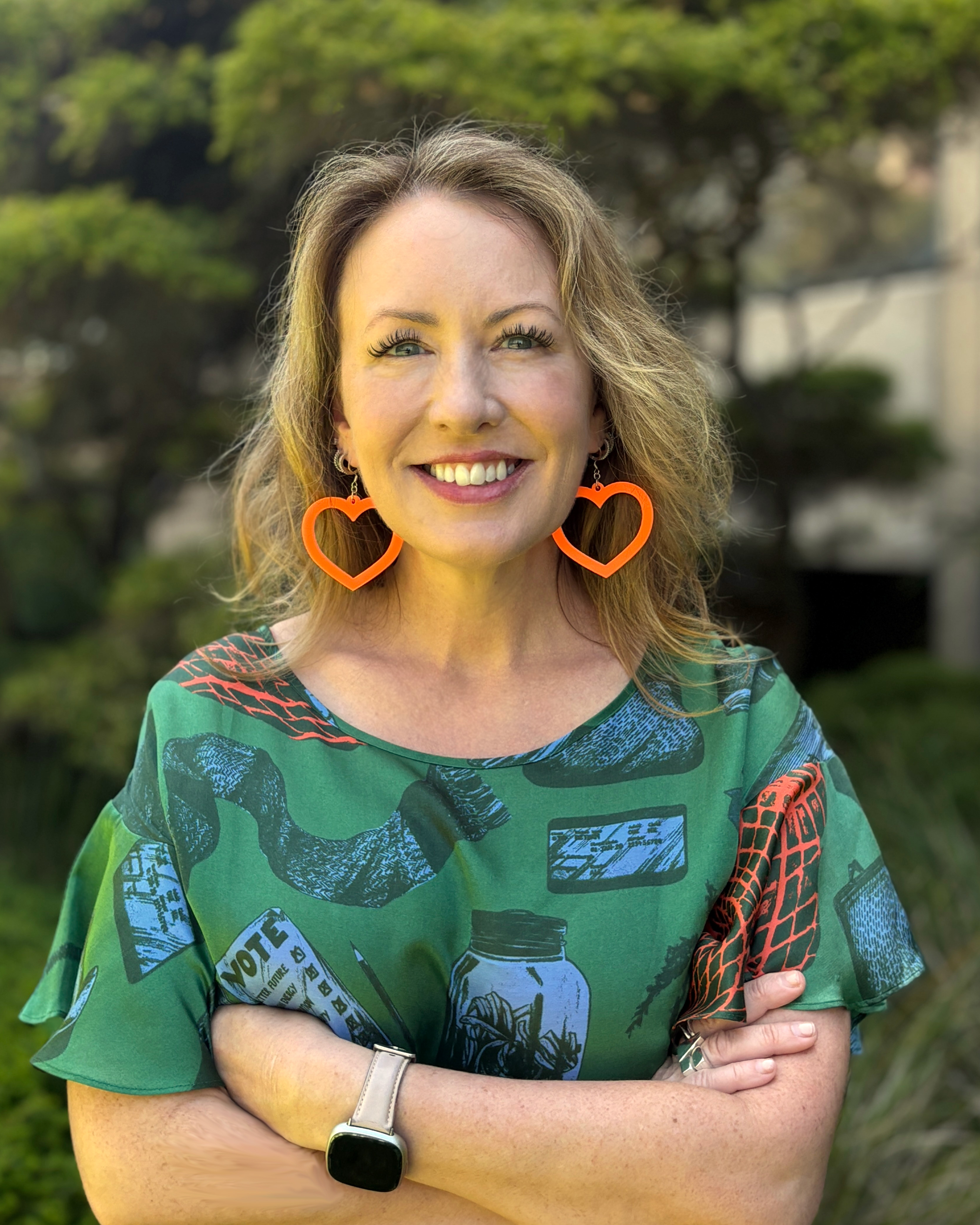 Abgail is wearing a green patterned top, smiling at the camera and is situated outside near a building with a tree in front of it