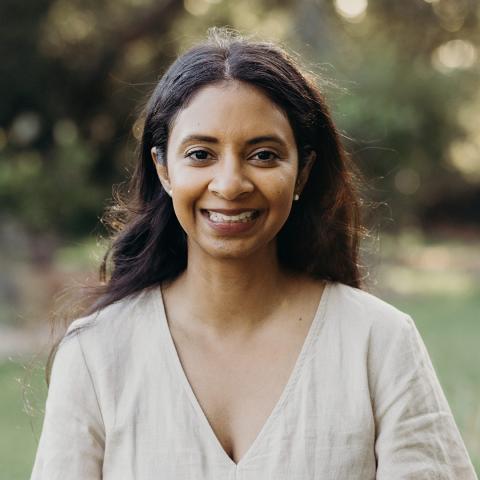 This is an image of Lini, a warmly smiling person in a beige coloured linen top, standing outdoors. She has long dark hair that is tucked behind her ears, where you can see she has small earring studs.