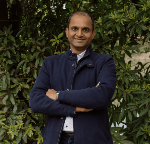 This is an image of Binoy, a smiling individual with a dark blue coat, a light blue collared shirt and denim jeans. He is standing outdoors, in front of dark green trees.