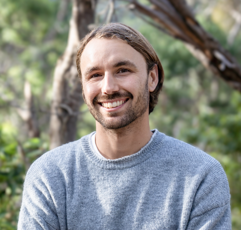 This is an image of Mitch, a smiling individual. Mitch has brown hair and is wearing a grey long sleeve shirt. He is standing outside in front of green trees on a sunny day. 