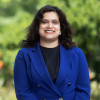 This is an image of Payal, a smiling individual with black hair. Payal is standing outdoors in front of light green trees, and is wearing a blue blazer over a black top. 