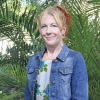 This is an image of Lauren, a smiling individual who is wear a light blue denim jacket and standing in front of green shrubbery on a sunny day.
