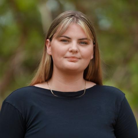 This is an image of Alyssa, who is standing outdoors in a blurred parkland environment. She has long brown hair with blond highlights and a fringe that frames her forehead. She's wearing a long sleeve black blouse with a small gold chain around her neck.