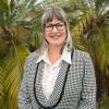 A woman with shoulder-length hair, wearing a stripy blazer and white button up shirt. Standing in front of a tree, smiling.