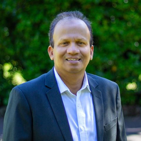 A man named Rifai, wearing glasses and a navy blazer over a white collared shirt, stands smiling against a backdrop of lush green foliage. The natural setting and his relaxed demeanor convey warmth and professionalism.