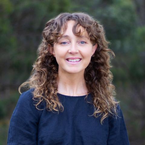 This is an image of Sophie, a brightly smiling individual standing in a bushland environment. She has brown curly hair with a fringe that sits just below her shoulders and wears a navy shirt. The background is blurred dark green, creating a calm and pleasant setting.