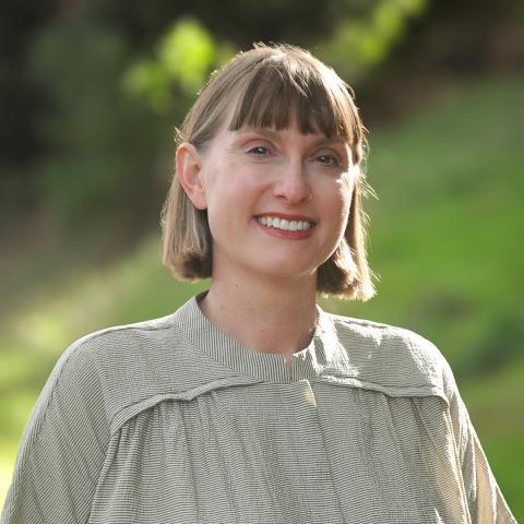 This is an image of Rachel, she's smiling warmly at the camera. She has light brown hair cut into a bob with bangs, and wears a white and green striped shirt with buttons down the middle. She's standing in front of a bright, green park landscape and the sun is shining creating a glow around Rachel.