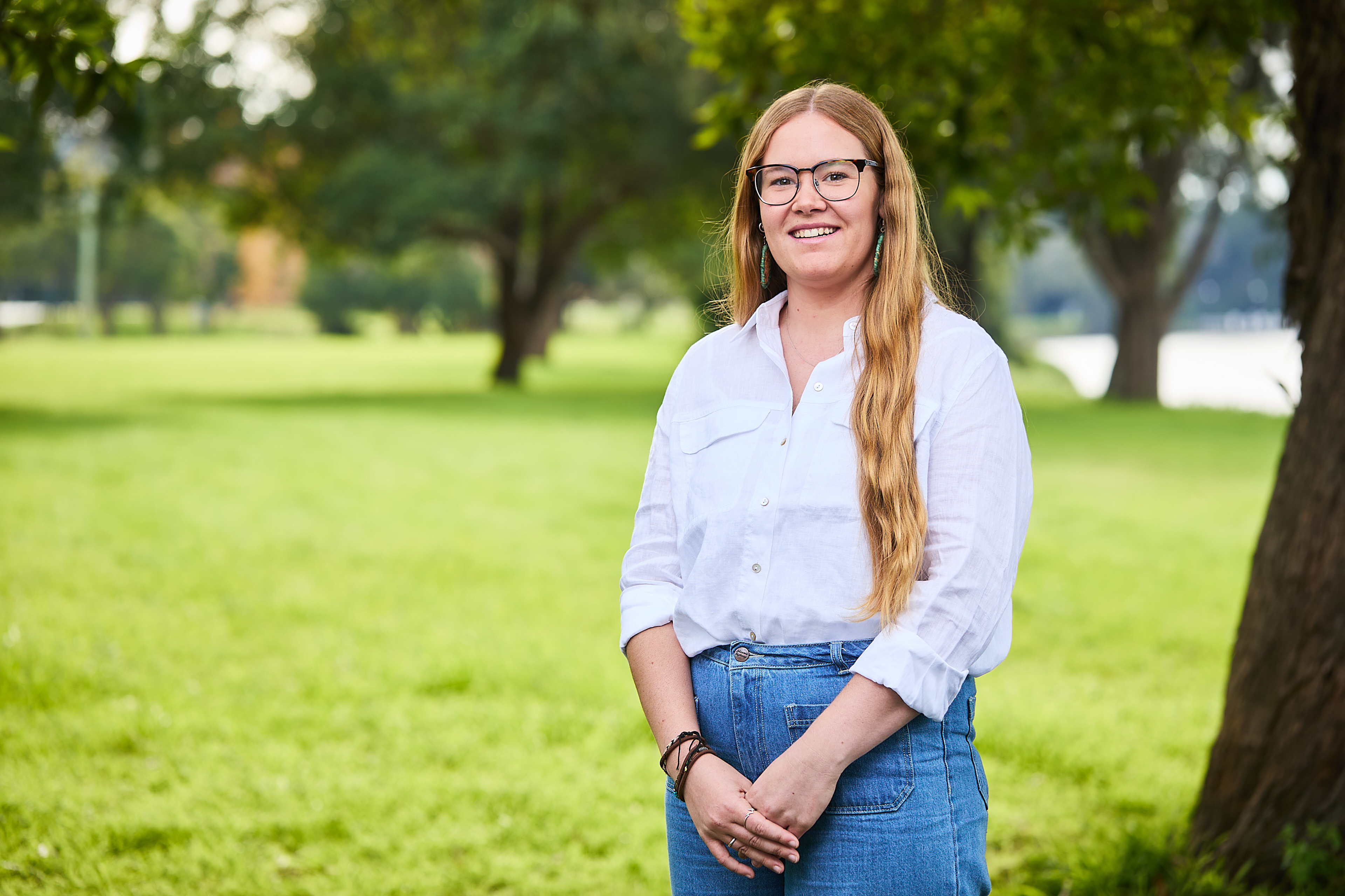 Takesa is wearing a white shirt and jeans, smiling at the camera and is situated on a grassy area outside with trees