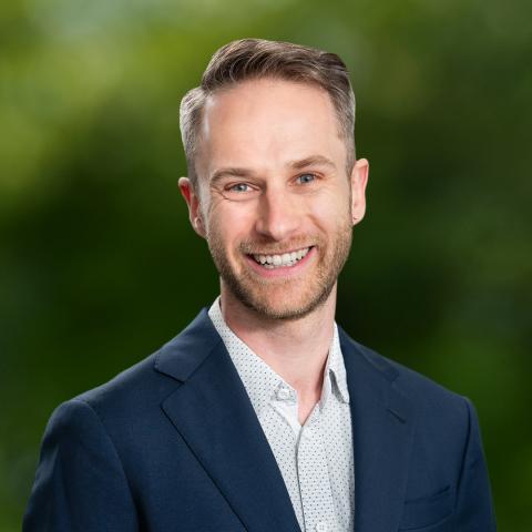 This is an image of Tim, who is warmly smiling at the camera. He is in a white collared shirt with small polka dots, and a navy blue blazer. His hair is fair and cut short, and he wears a small earring in his left ear.