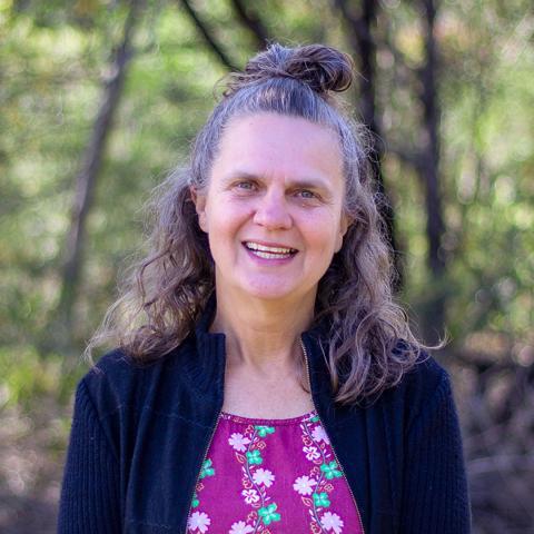 An image of Liza, a smiling woman standing outdoors in a bushland environment. She has silver-brown hair that sits just below her shoulders and half is tied up into a bun. She's wearing a pink, floral top and a navy cardigan and smiles warmly at the camera.