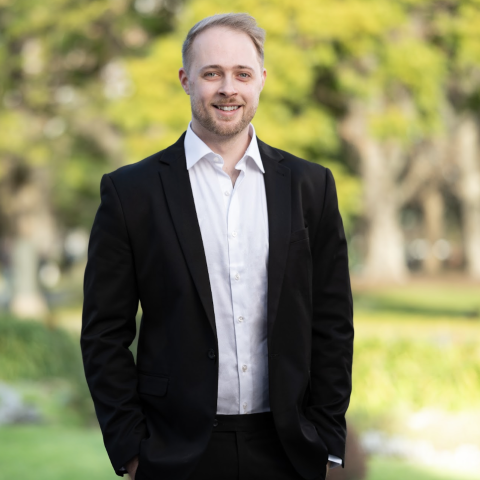 This is an image of Tomas, a smiling individual wearing a black suit and white button down collared shirt underneath. He is standing outside, in front of light green trees and grassy grounds.  