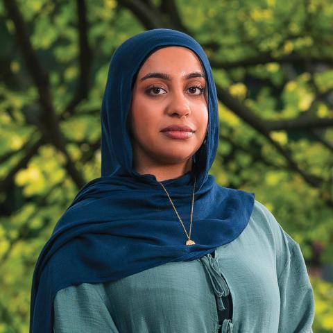 This is an image of Maryam, a smiling individual who has brown skin, and is wearing a dark blue hijab and a teal blue dress. Maryam is standing in front of light green trees on a sunny day.
