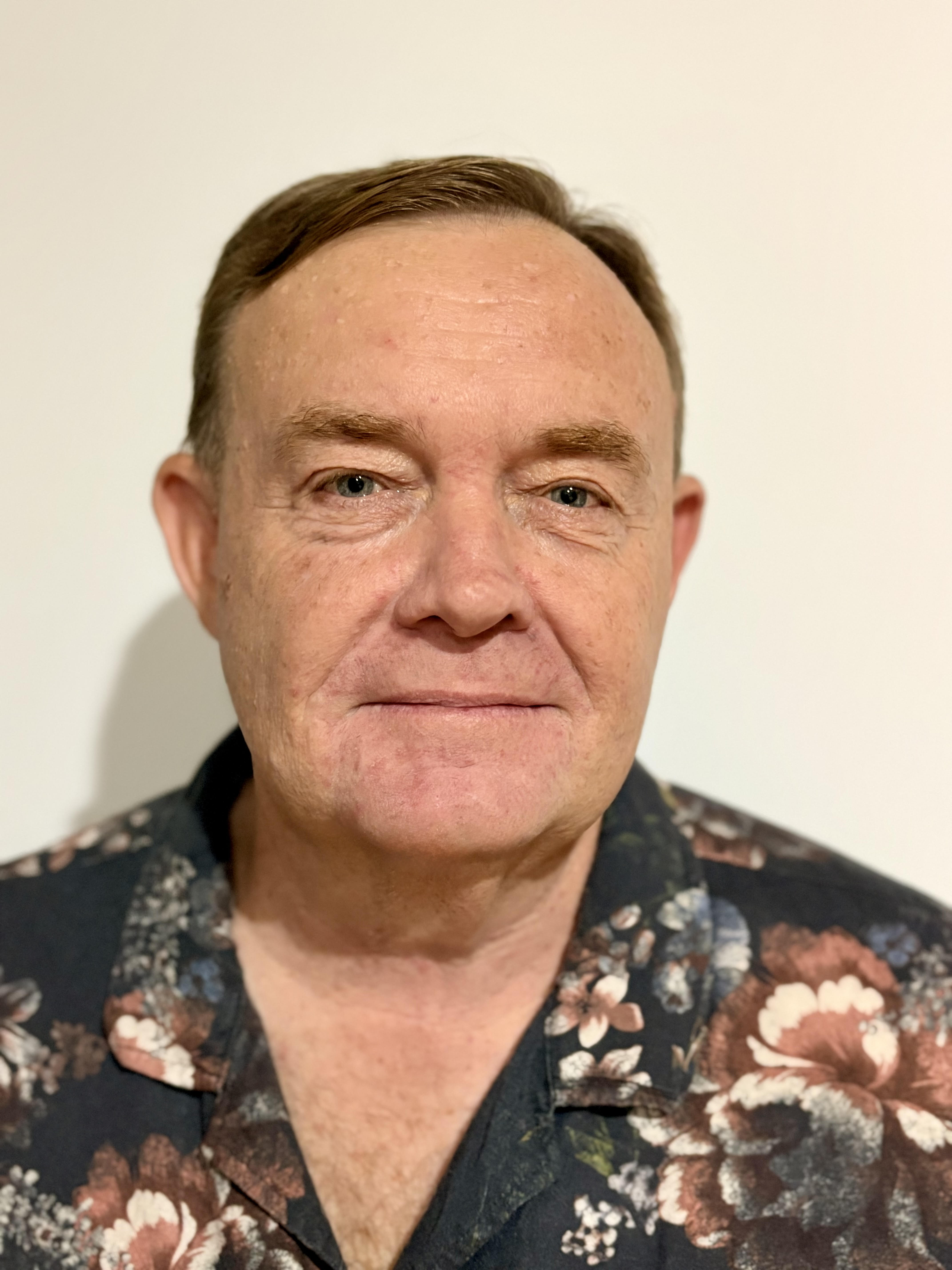 Grant is wearing a dark shirt with a pattern, smiling at the camera and situated in front of a light coloured wall