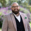 This is an image of Declan, a smiling individual with glasses. Declan is standing outdoors, in front of a colourful garden and wearing a beige blazer, black vest and white collared shirt. 