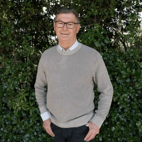 This is an image of Dave, a smiling individual. Dave is wearing glasses, a pullover and a collared shirt. He is standing in front of dark green trees on a sunny day.