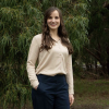 This is an image of Jill, a smiling individual with long, dark hair. She is standing outside, in front of dark green trees and is wearing a beige top and black pants.