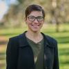 A woman with short brown hair and glasses, wearing a dark green shirt and blazer standing in a park smiling
