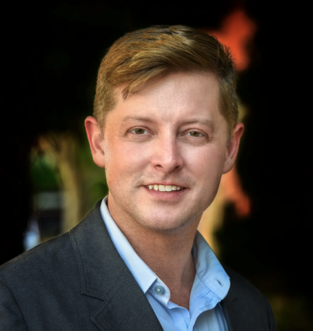 This is an image of Paul, a smiling individual with brown hair and a blue collared shirt, and a blazer on top.