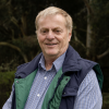 This is an image of Jeff, a smiling individual with a collared striped shirt and a black and green vest. Jeff is standing outside in front of dark green trees.
