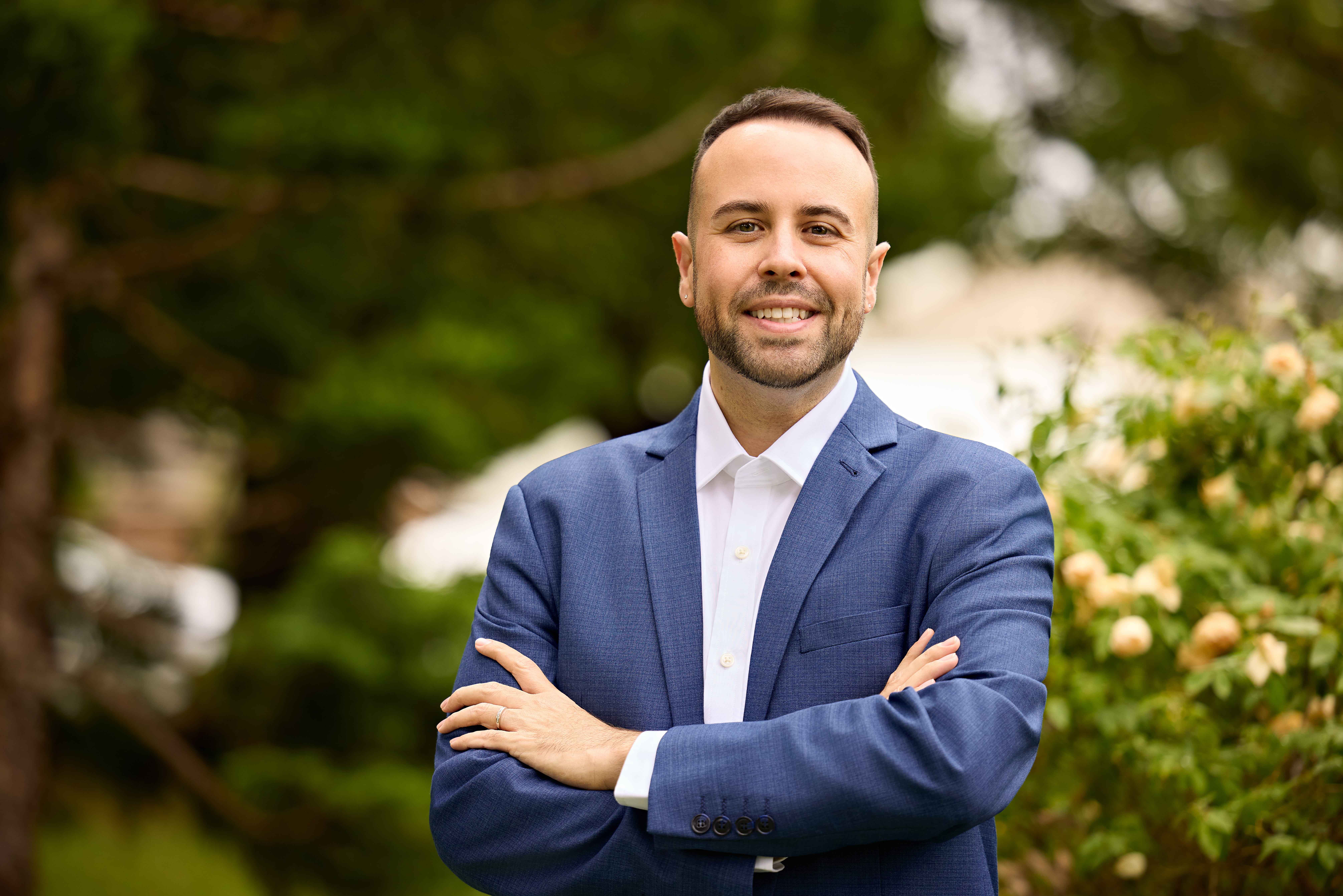 Shane is wearing a suit, smiling at the camera and situated outside in front of greenery