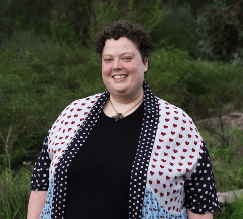This is an image of Maggie, a smiling individual with short dark hair. Maggie is standing outside, in front of dark green bushes and wearing a white, red and black blouse. 