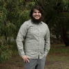 This is an image of Timon, a smiling individual with dark hair and a dark beard, wearing a green pinstriped button down. He is standing outside, in front of lush green trees.