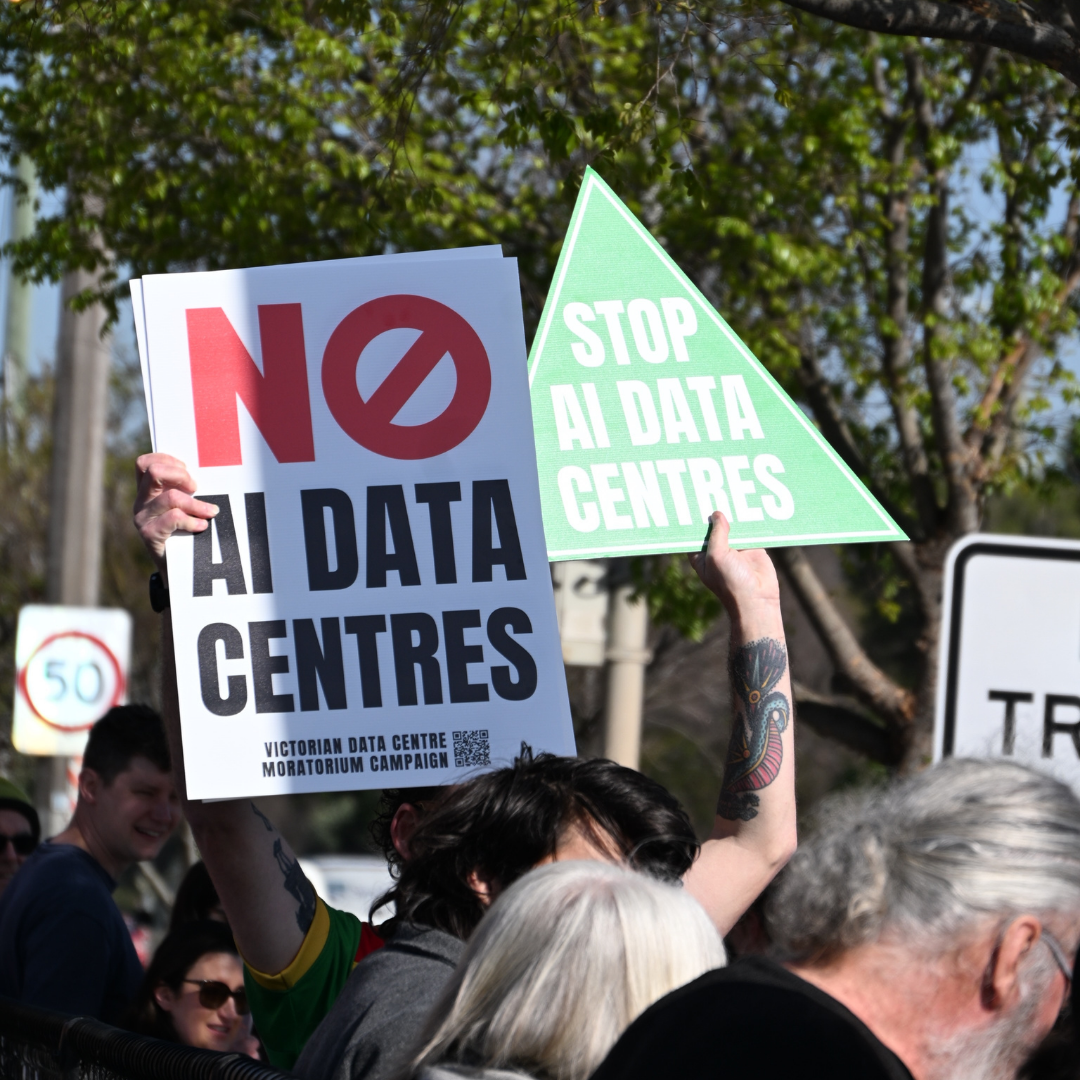 Signs from AI data centre protests in Melbourne. Greens triangle reads Stop AI Data Centres, White sign reads No AI Data Centres