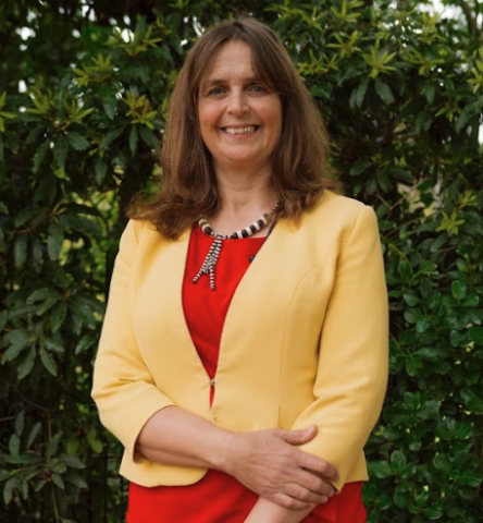 This is an image of Catherine, a smiling individual standing in front of dark green leaves. Catherine is smiling warmly and wearing a yellow blazer on top of a red blouse.