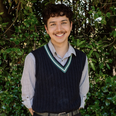 This is an image of Kai, a smiling individual standing outside, in front of dark green trees. Kai is wearing a collared white and blue striped shirt with a dark blue vest pullover on top.