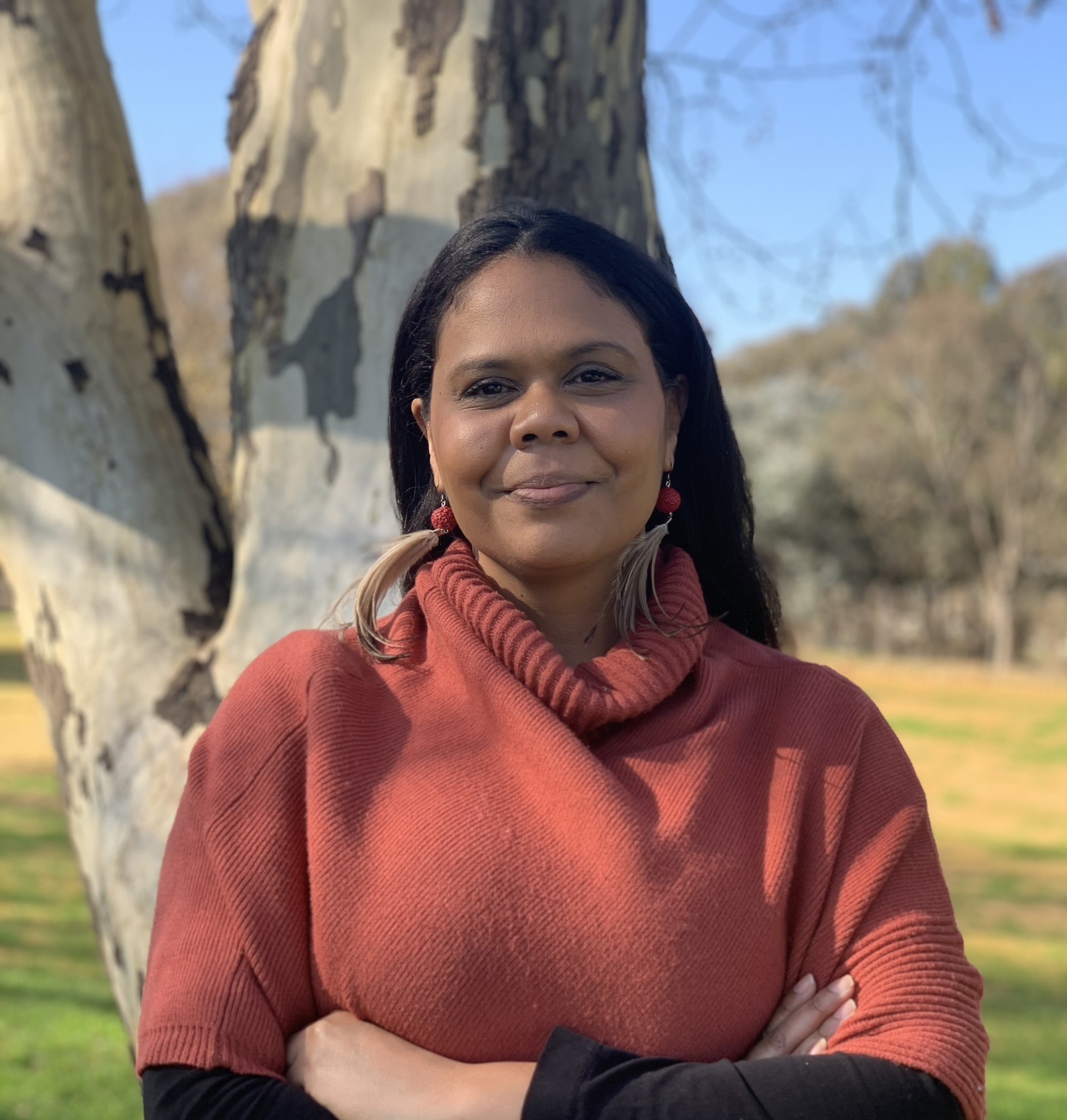 Lynda-June is wearing an orangy red jumper, smiling at the camera and is situated outside in front of a tree