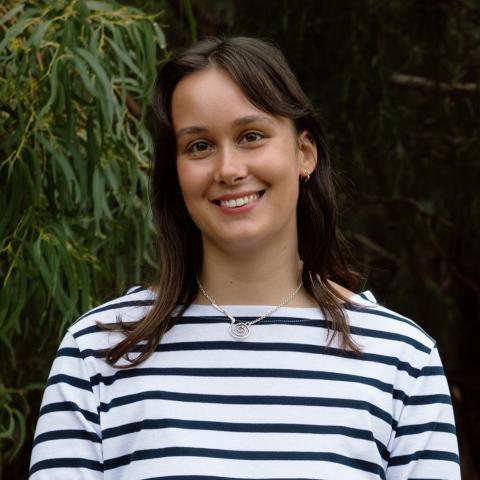This is an image of Katie, a warmly smiling woman in an outdoor environment. Katie has brown, straight hair that falls just below her shoulders with a side fringe that's tucked behind her left ear. She wears a boat neck white top with navy stripes and a small pendant around her neck.