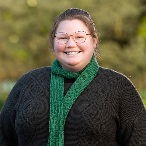 This is an image of Zoe, a brightly smiling person standing outdoors. She has dark blonde hair tied up into a ponytail and round glasses. She's wearing a dark knit top and a forest green scarf around her neck.