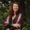 This is an image of Karli, a smiling individual with dark red hair. Karli is standing outside, in front of green trees and bushes. She is wearing a moron blazer over a black top, with a colourful necklace and bracelets. 