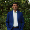 This is an image of Roshan, a smiling individual. Roshan is wearing a dark blue suit with a light blue collared button down shirt. He is standing in front of green bushes.