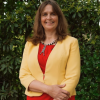 This is an image of Catherine, a smiling individual standing in front of dark green leaves. Catherine is smiling warmly and wearing a yellow blazer on top of a red blouse.